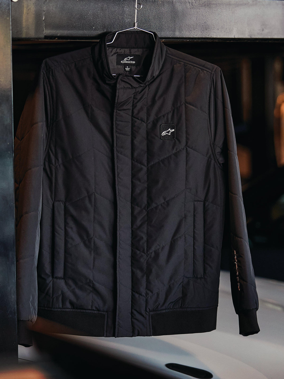 Alpinestars Precedent Jacke, gesteppte Sportswear-Jacke, Schwarz, mit schrägem Steppmuster, niedrigem Stehkragen mit verdecktem Reißverschluss, Logo-Patch auf der Brust und Rippbündchen an Ärmeln und Saum, Motorsport-inspiriertes Design