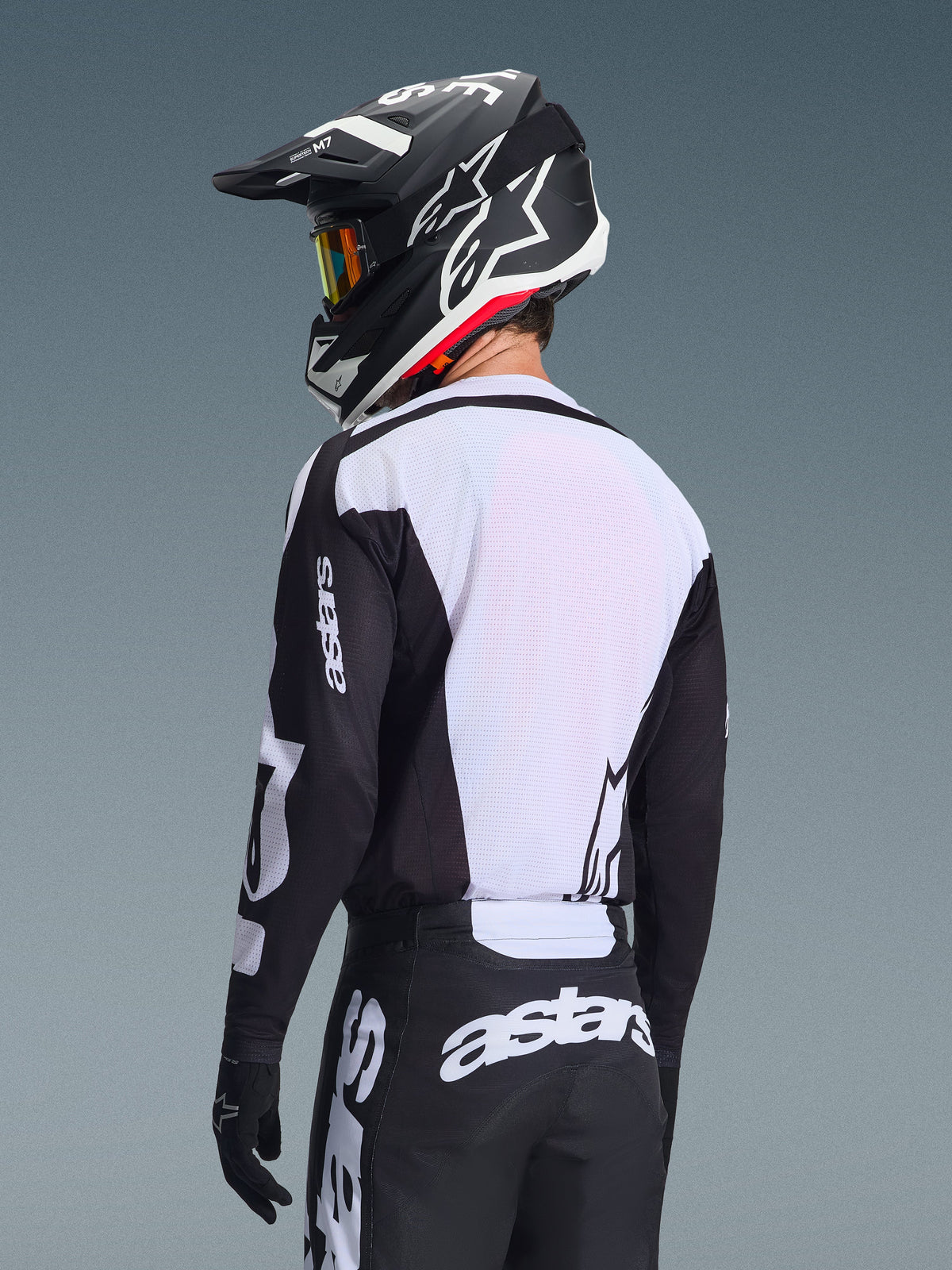 Pilote de motocross de dos portant le maillot Alpinestars 2026 Racer Air Riway, noir et blanc, doté d'un panneau dorsal en mesh blanc respirant et de manches noires avec marquage 'astars', pantalon noir assorti avec logos blancs, casque noir avec accents blancs, équipement tout-terrain professionnel pour des performances par forte chaleur.