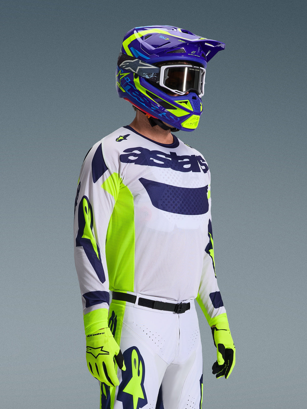 Motocross-Fahrer im Alpinestars 2026 Racer Air Riway Bekleidungsset, weißes Jersey mit marineblauem Logo und neongelben Seiteneinsätzen, weiße Hose mit passenden marineblauen und gelben Akzenten, lila und neongelber Helm mit weißer Brille, neongelbe Handschuhe, Hochleistungs-Offroad-Rennausrüstung