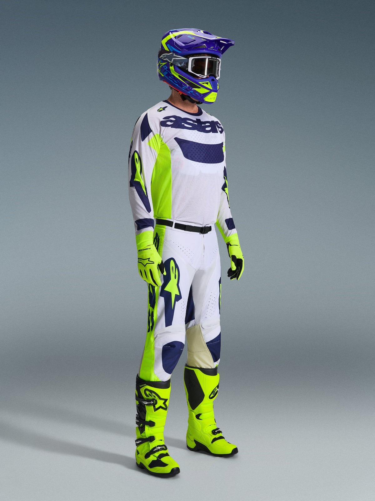 Motocross-Fahrer im Alpinestars 2026 Racer Air Riway Gear Set in Hellgrau, Neongelb und Blau. Das Set umfasst ein langärmeliges Mesh-Trikot, eine passende Rennhose, leuchtend neongelbe Handschuhen und Stiefel sowie einen lila-blauen Helm mit Schutzbrille, entwickelt für belüftete Offroad-Performance.