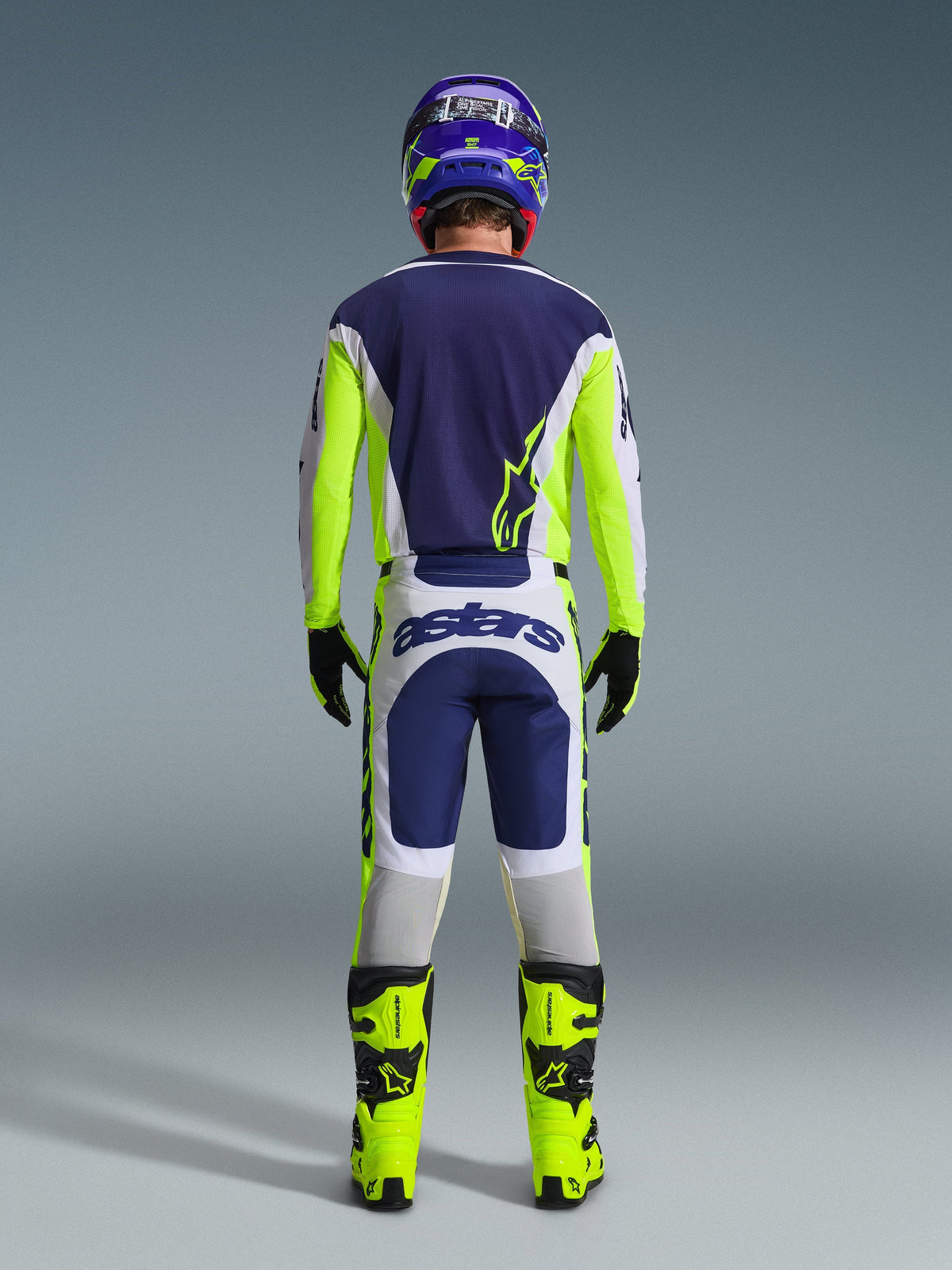 Motocross-Fahrer von hinten im Alpinestars 2026 Racer Air Riway Gear Set, blaues Trikot mit neongelben und hellgrauen Akzenten, weiß-blaue Hose mit neongelben Seiteneinsätzen, neongelbe Stiefel mit schwarzen Details, passender blau-gelber Helm, professionelle Ausrüstung für Offroad-Fahrten bei warmem Wetter
