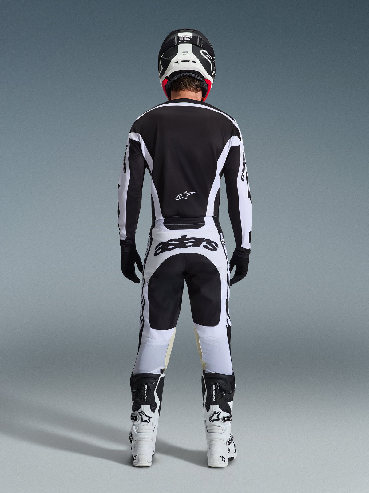 Vue arrière d'un pilote de motocross portant l'ensemble d'équipement Alpinestars Racer Riway 2026 en noir et blanc, comprenant un maillot noir avec des accents blancs, un pantalon blanc avec le logo « astars » sur l'assise, un casque noir, des bottes blanches et des gants noirs, tenue de course tout-terrain professionnelle complète.