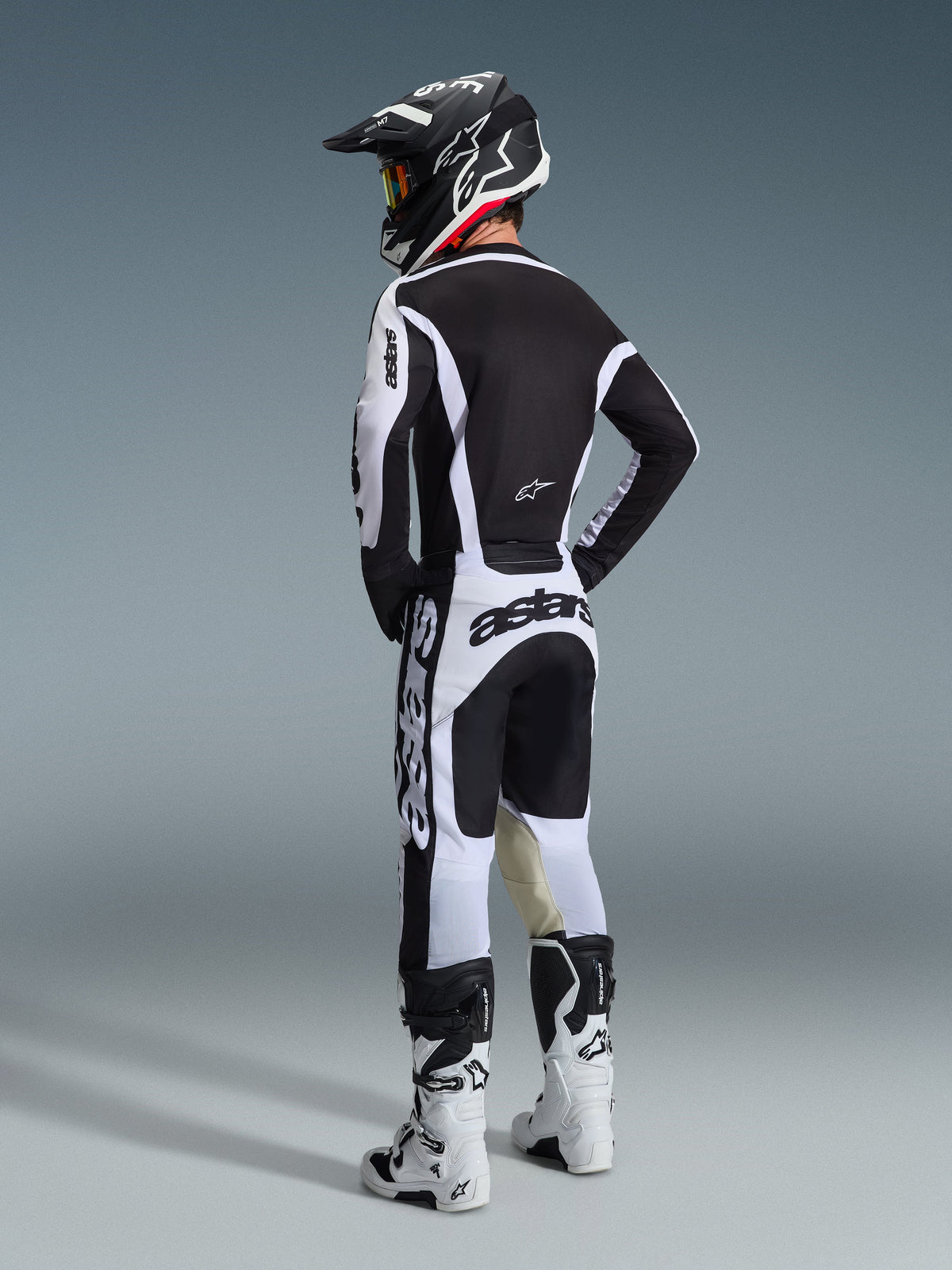 Pilote de motocross vu de dos, portant l'ensemble d'équipement Alpinestars Racer Riway 2026 noir et blanc, avec un casque noir assorti et des bottes de motocross blanches, design épuré haute performance pour le tout-terrain