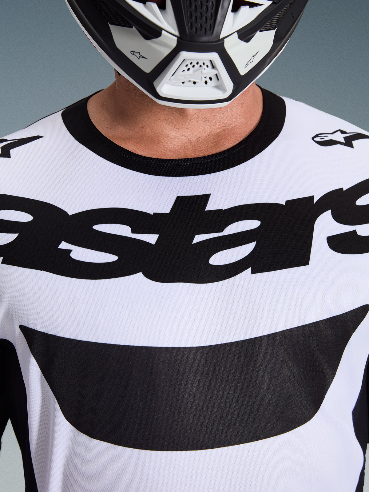 Gros plan d'un pilote portant le maillot Alpinestars 2026 Racer Riway en blanc et noir, arborant un logo 'astars' audacieux sur la poitrine et un col rond noir, avec un casque de motocross assorti.
