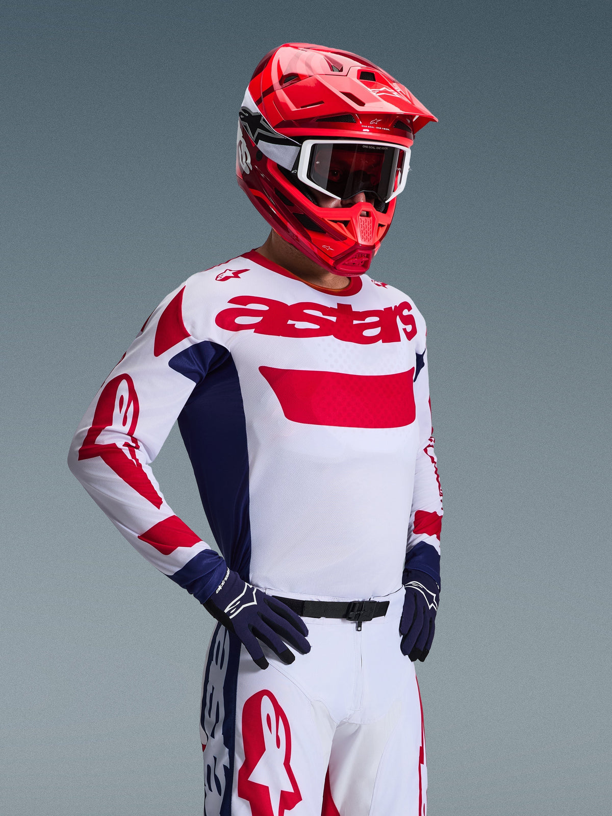 Pilote de motocross portant le maillot Alpinestars 2026 Racer Riway en blanc, bleu et rouge, pantalon tout-terrain assorti, casque rouge vif avec lunettes blanches et gants bleu marine, ensemble d'équipement haute performance coordonné pour le motocross