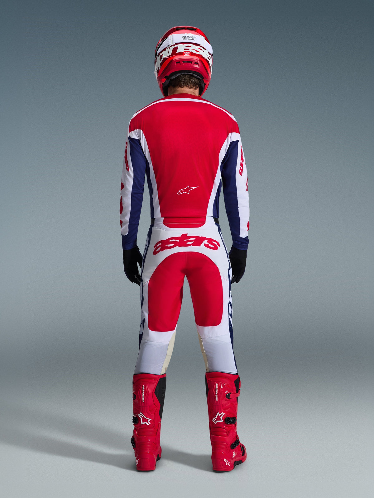 Pilote de motocross vu de dos portant l'ensemble d'équipement Racer Riway 2026, maillot rouge et bleu avec accents blancs, pantalon blanc avec empiècements rouges et logo 'astars' sur l'assise, coordonné avec un casque rouge, des bottes rouges et des gants noirs, équipement de course haute performance pour le tout-terrain