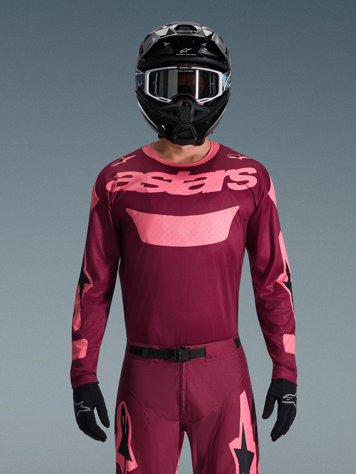 Pilote de motocross portant l'ensemble Alpinestars 2026 Racer Riway, bordeaux et rose, maillot avec marquage rose imposant sur la poitrine, pantalon assorti avec accents noirs et roses, casque intégral noir avec lunettes, et gants noirs, tenue de course tout-terrain coordonnée.