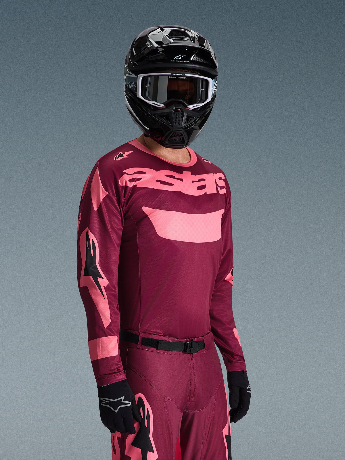 Pilote de motocross portant l'ensemble Alpinestars 2026 Racer Riway, maillot bordeaux et rose avec marquage 'astars' imposant, pantalon assorti avec accents roses, casque et gants noirs, tenue de course professionnelle pour motocross tout-terrain