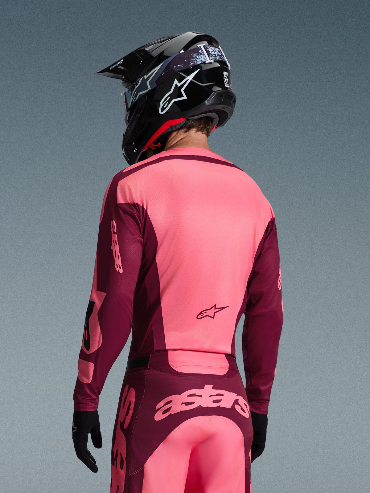 Pilote de motocross vu de dos portant l'ensemble d'équipement Alpinestars Racer Riway 2026, maillot et pantalon rose vif et bordeaux, casque de motocross noir avec logo blanc et gants noirs, arborant le marquage « astars » dans le dos.