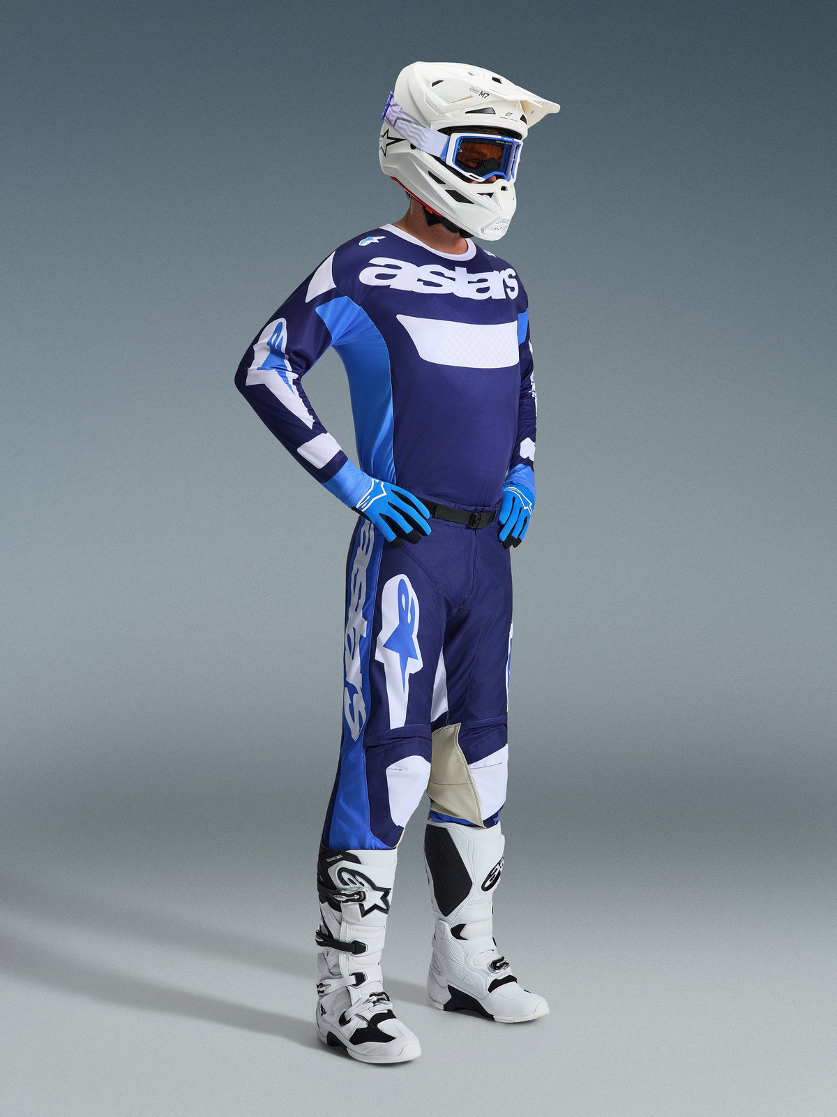 Pilote de motocross portant l'ensemble d'équipement Alpinestars Racer Riway 2026 en bleu UCLA et blanc, comprenant un maillot bleu et blanc avec logo 'astars', un pantalon assorti, un casque blanc avec lunettes bleues et des bottes tout-terrain blanches, tenue de course complète pour le motocross.