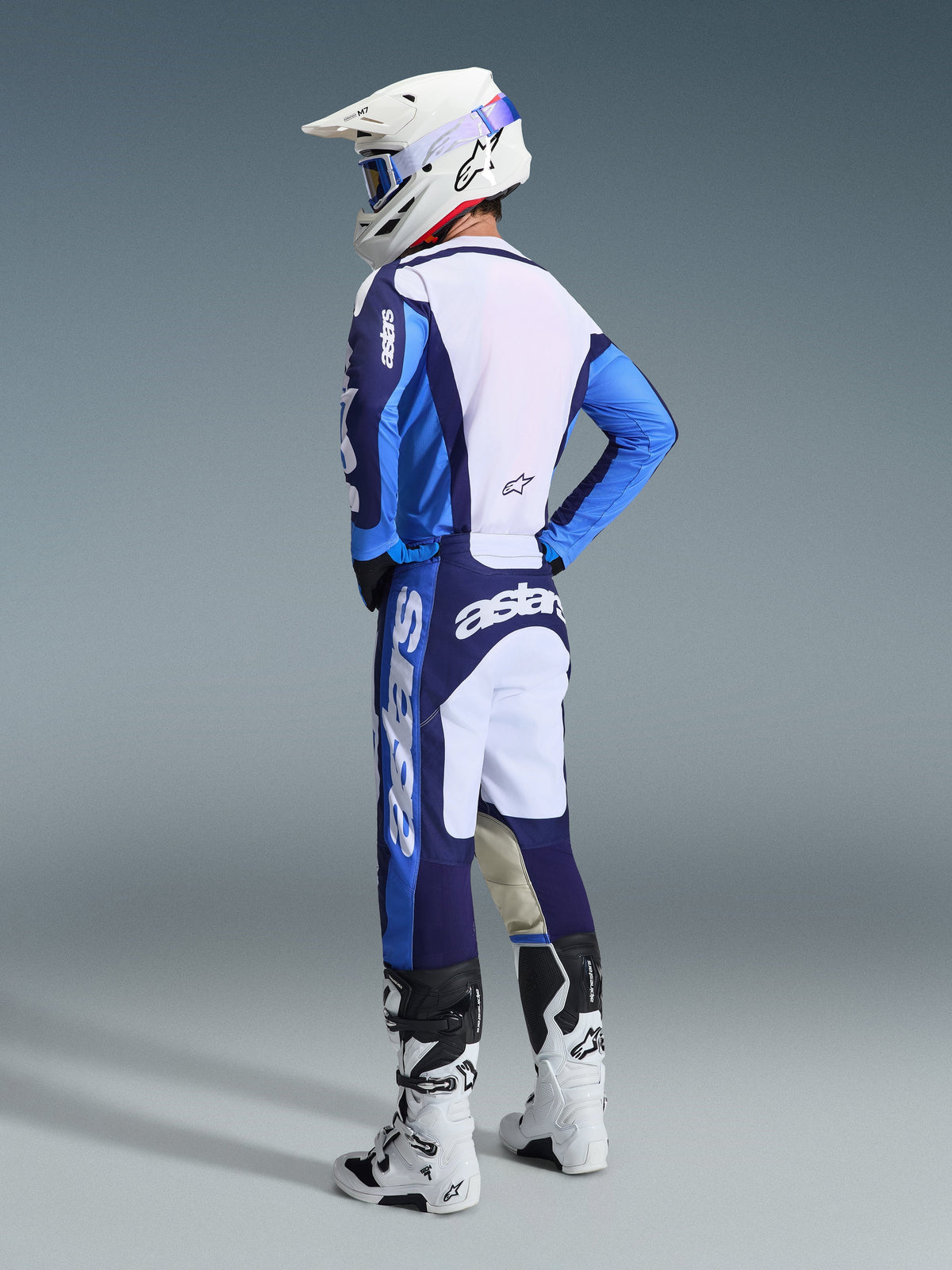 Pilote de motocross vu de trois-quarts arrière portant l'ensemble Alpinestars Racer Riway 2026 en bleu UCLA et blanc, composé d'un maillot bleu et blanc avec marquage « astars », d'un pantalon assorti, d'un casque de motocross blanc avec lunettes bleues et de bottes de la série Tech blanches et noires.