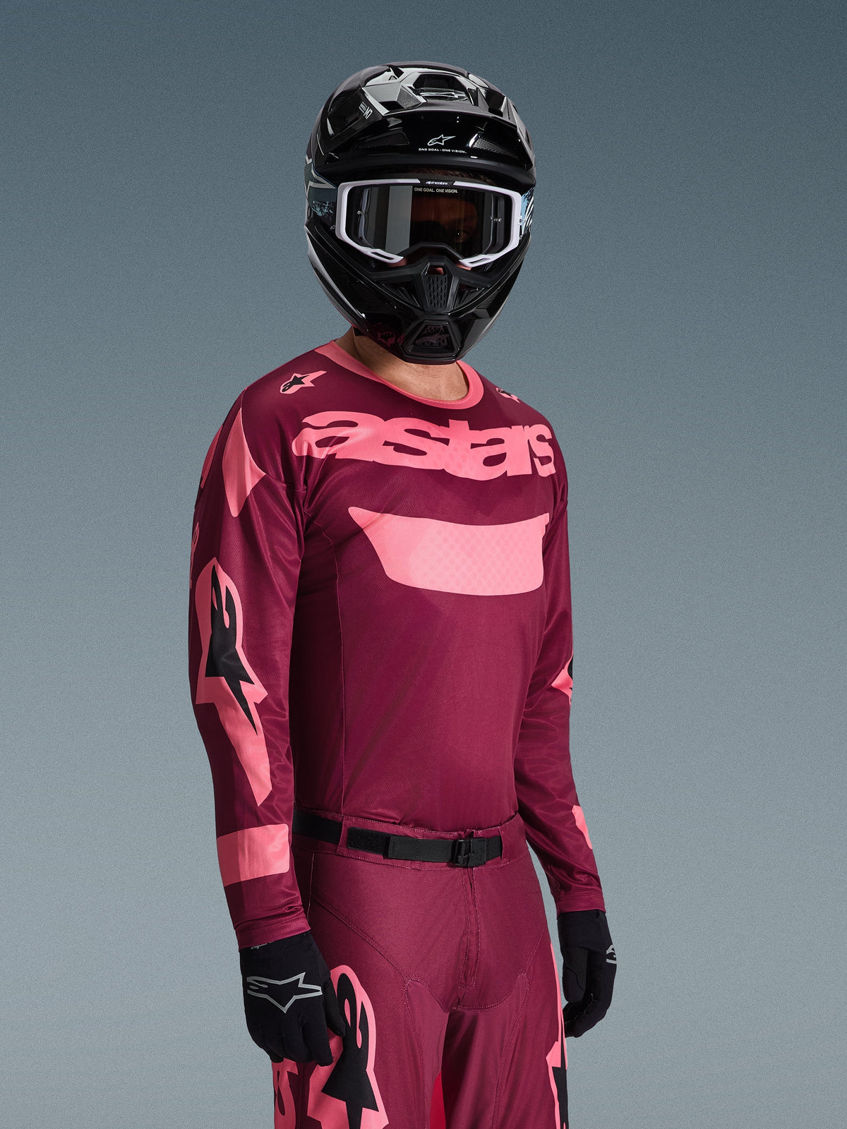 Motocross-Fahrer im Alpinestars 2026 Racer Riway Trikot in Dark Red Coral mit passender Hose, korallenrosa „astars“-Branding auf der Brust, dazu ein schwarzer Helm und schwarze Handschuhe, abgestimmtes Offroad-Rennausrüstungsset.