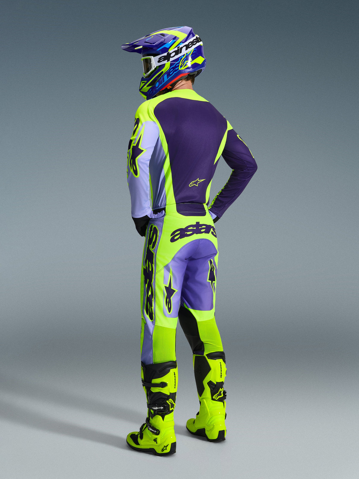 Pilote de motocross portant l'ensemble d'équipement Alpinestars 2026 Racer Portl, maillot et pantalon violets et jaune fluo avec empiècements lavande, casque assorti violet et jaune, bottes jaune fluo éclatant, vue arrière montrant l'équipement de course tout-terrain professionnel.