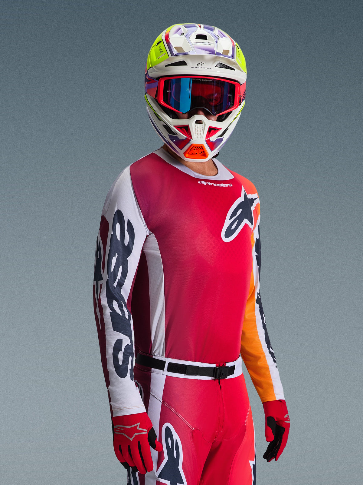 Motocross-Fahrer im Alpinestars 2026 Racer Portl Gear Set, mit einem roten Jersey mit Pink-Tönen und weißen Ärmeln, passender roter Hose, weißem Helm mit leuchtend gelben und lila Akzenten, roter Brille mit blau schillernden Gläsern und roten Handschuhen, professionelle Rennausrüstung für Motocross.