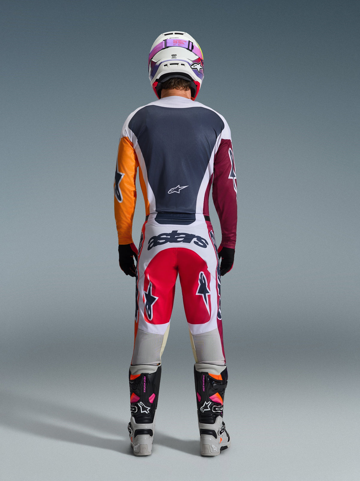 Rückansicht eines Motocross-Fahrers im Alpinestars 2026 Racer Portl Gear Set, mehrfarbiges Trikot mit orangefarbenen und weinroten Ärmeln, weiß-rote Hose mit „astars“-Branding am Gesäß, passender weißer Helm und technische Rennstiefel, entwickelt für Offroad-Motocross-Performance.