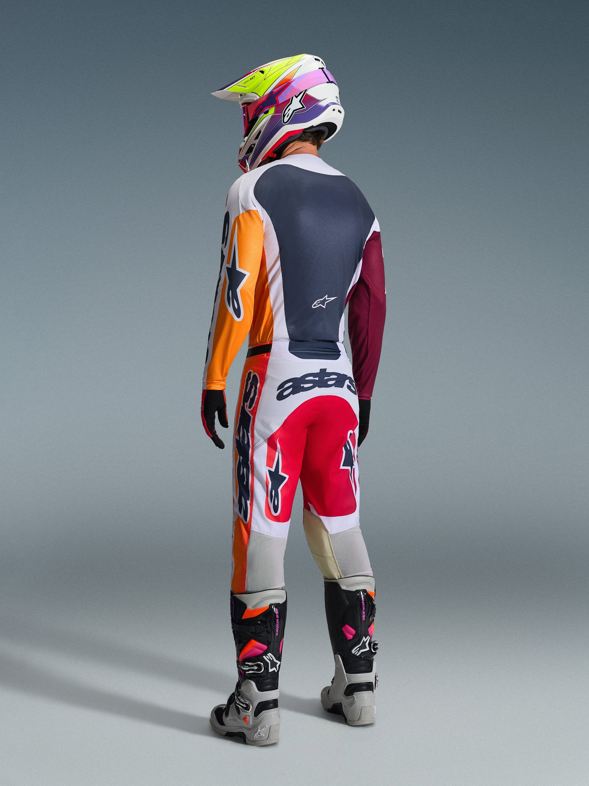 Alpinestars 2026 Racer Portl Gear Set, Motocross-Fahrer in der Rückansicht, Trikot im Color-Block-Design mit orangefarbenen und kastanienbraunen Ärmeln, weiße Hose mit roten Einsätzen und markantem „astars“-Branding, mehrfarbiger Helm und graue Stiefel, professionelles Offroad-Rennoutfit.