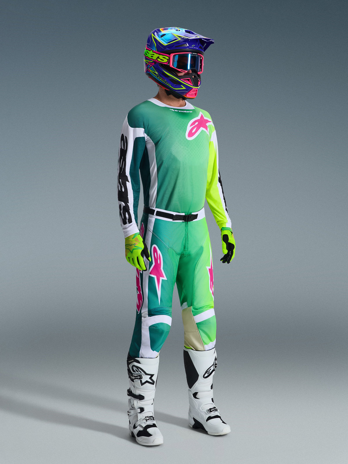 Alpinestars 2026 Racer Portl Gear Set, Motocross-Fahrer in einem Jersey mit Farbverlauf von Blaugrün zu Limettengrün, pinkfarbene Logos und weiße Ärmel, passende grün-weiße Hose, bunter Grafikhelm mit pinker Brille, weiße Stiefel und fluoreszierend gelbe Handschuhe, professionelle Offroad-Rennausrüstung.