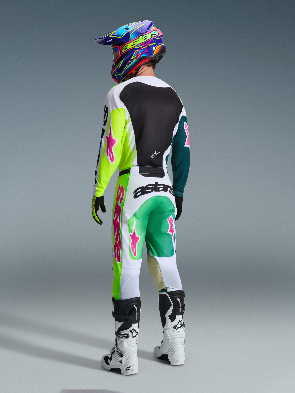 Motocross-Fahrer von hinten im Alpinestars 2026 Racer Portl Gear Set, Jersey mit neongelben und türkisblauen Ärmeln sowie pinken Logos, passende weiß-grüne Hose mit auffälligen pinken Akzenten, bunter Helm und weiße Stiefel, professionelle Offroad-Rennausrüstung