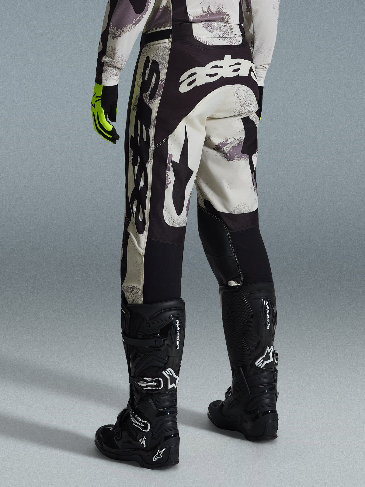 Alpinestars 2026 Racer Lahnd broek, motorcrossbroek, Sand Camo en grijs, achteraanzicht met opvallende witte branding op het zitvlak en de pijpen, gecombineerd met zwarte motorcrosslaarzen en gele handschoenen, professionele off-road race-uitrusting