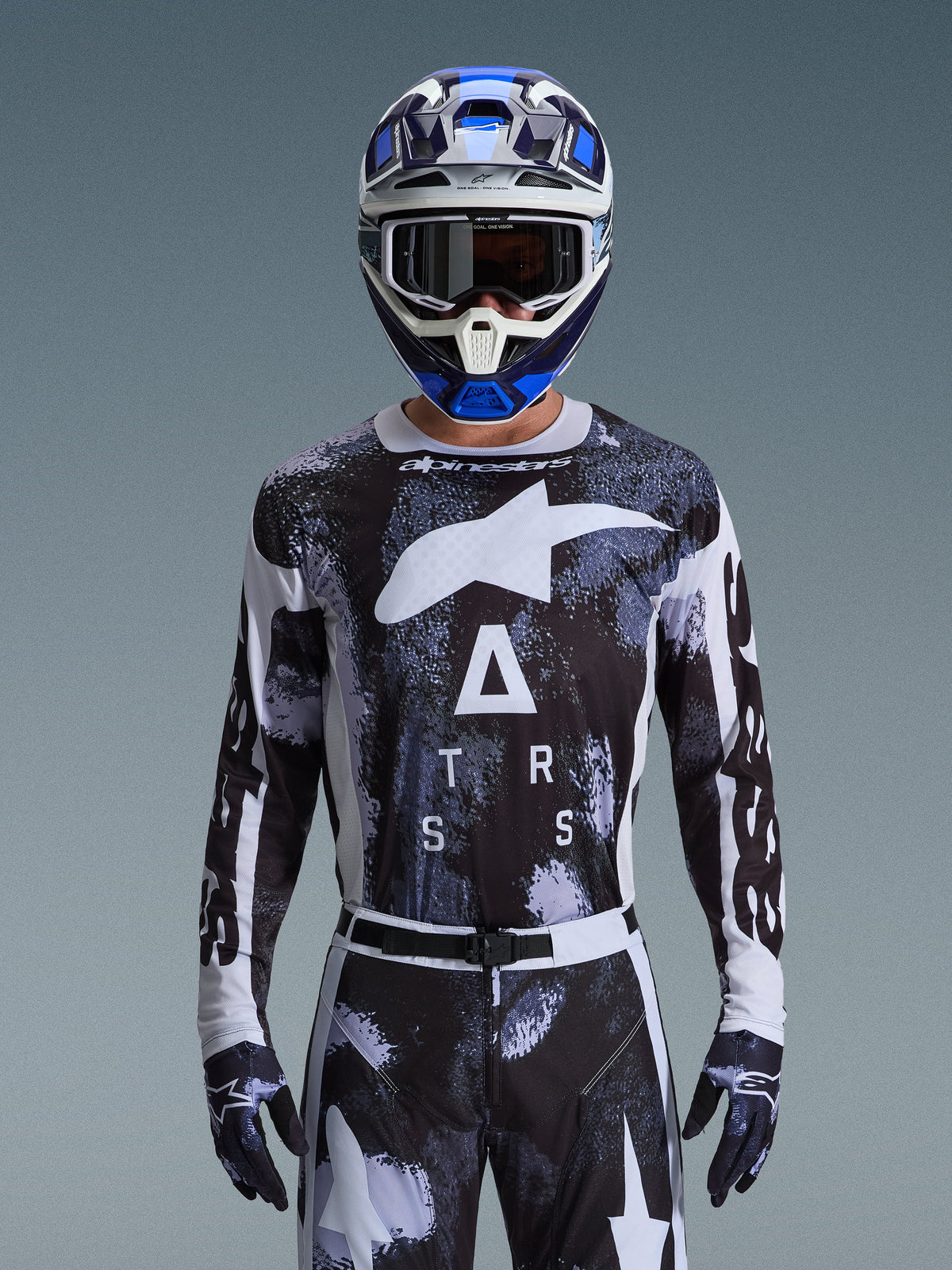 Alpinestars 2026 Racer Lahnd Gear Set, motocross-rijder in zwart-wit abstract camouflage shirt met grote witte logo's en "ATRS"-belettering, bijpassende broek en handschoenen, blauw-witte helm met bril, hoogwaardige race-outfit voor off-road motorcross