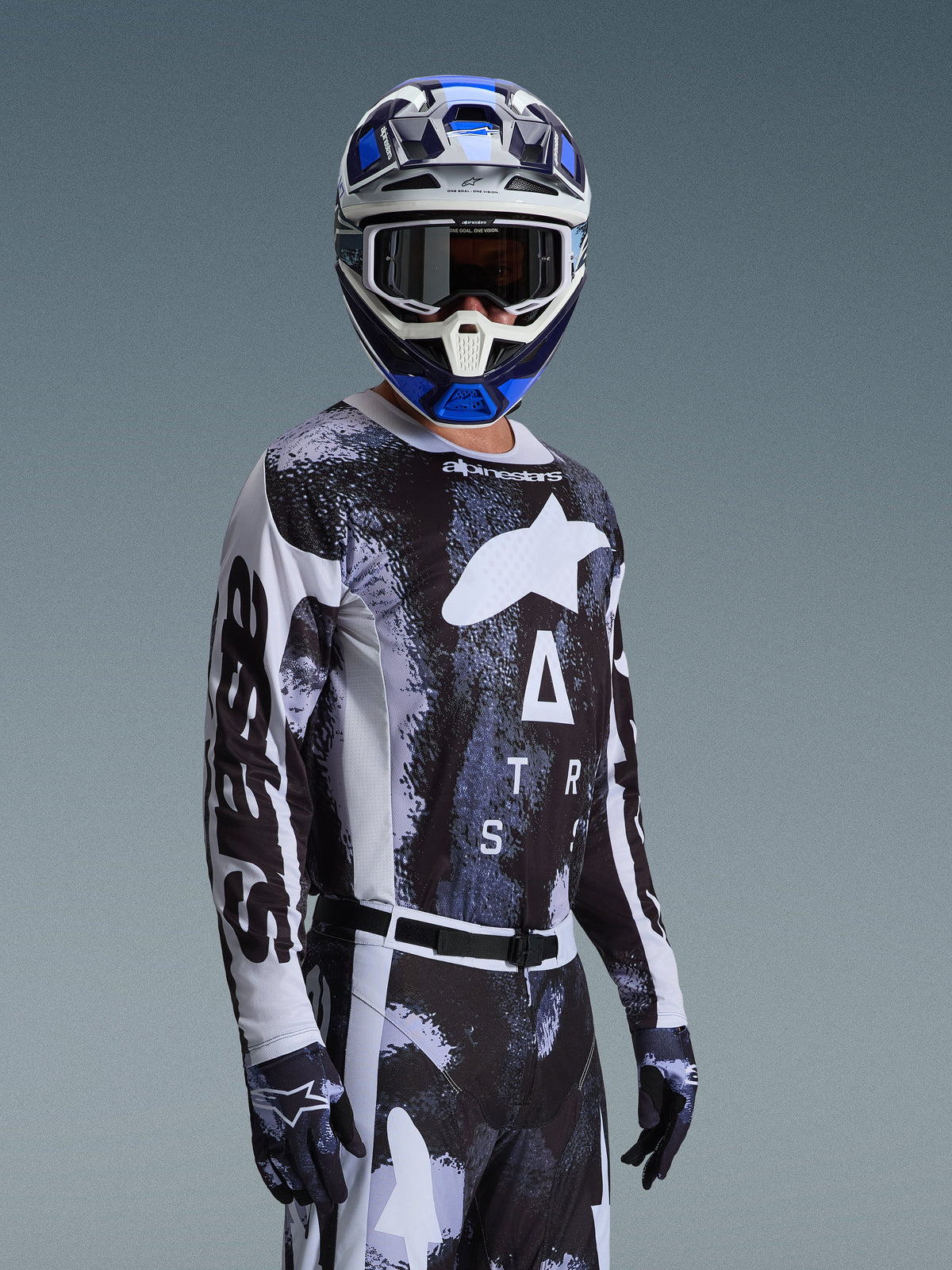 Motorcrossrijder in Alpinestars 2026 Racer Lahnd Gear Set, zwart-wit jersey met gemarmerde blauwe en grijze graphics, bijpassende broek en handschoenen, blauw-witte helm met crossbril, performance-uitrusting voor off-road racen