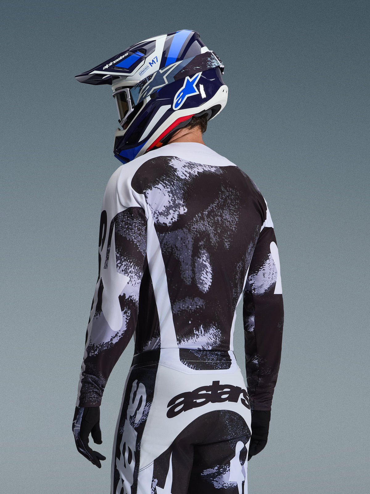 Alpinestars 2026 Racer Lahnd Gear Set, motorcrosser van achteren gezien in jersey en broek met zwart-witte gespikkelde graphics, gecombineerd met een blauw-witte Alpinestars helm en zwarte handschoenen, hoogwaardige off-road race-uitrusting