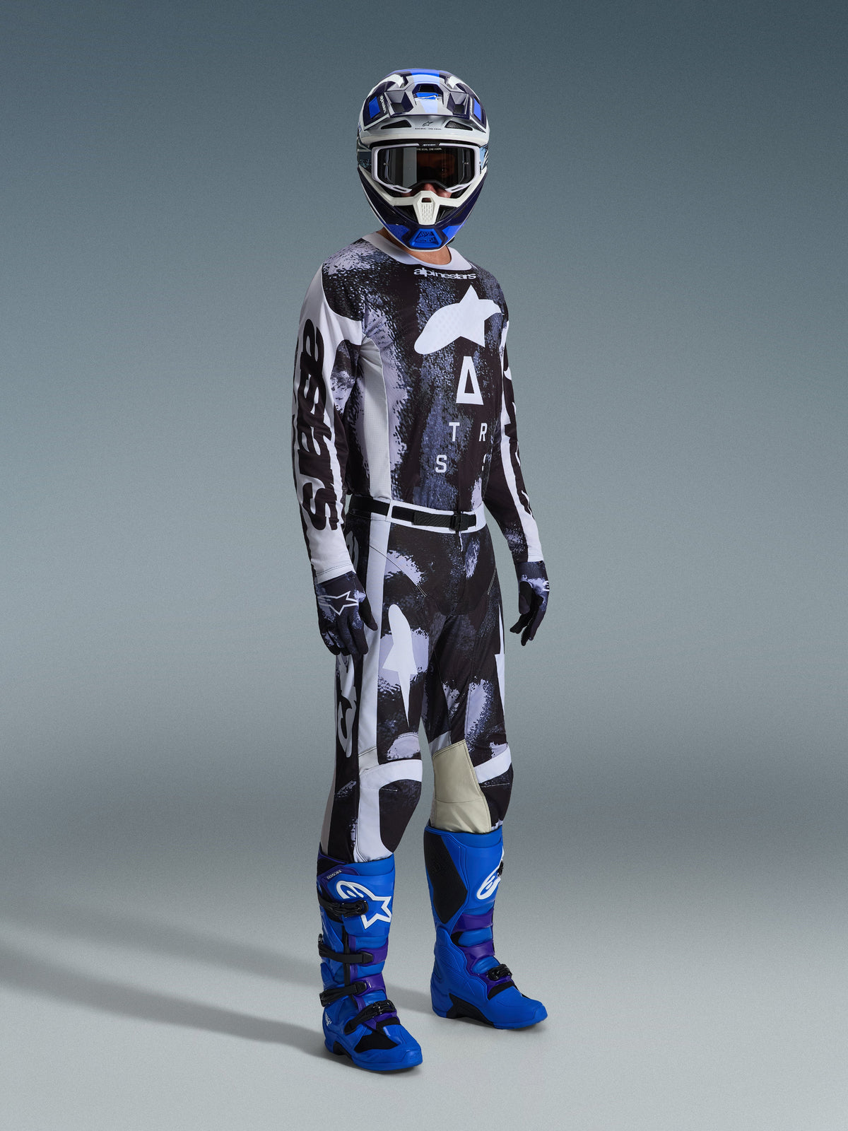 Alpinestars 2026 Racer Lahnd Gear Set, motocrossrijder met een zwart-wit bedrukt shirt en broek met een abstract verweerd ontwerp, voorzien van grote witte Alpinestars-logo's, gecombineerd met felblauwe en paarse laarzen en een blauw-witte helm, complete professionele off-road race-outfit.