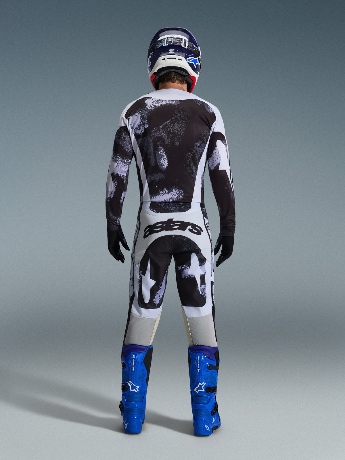 Motorcrossrijder van achteren gezien in de Alpinestars 2026 Racer Lahnd Gear Set, zwart-wit jersey en broek met abstract patroon, 'astars' branding op het zitvlak, gecombineerd met blauwe laarzen en een bijpassende helm, professionele off-road racekleding.