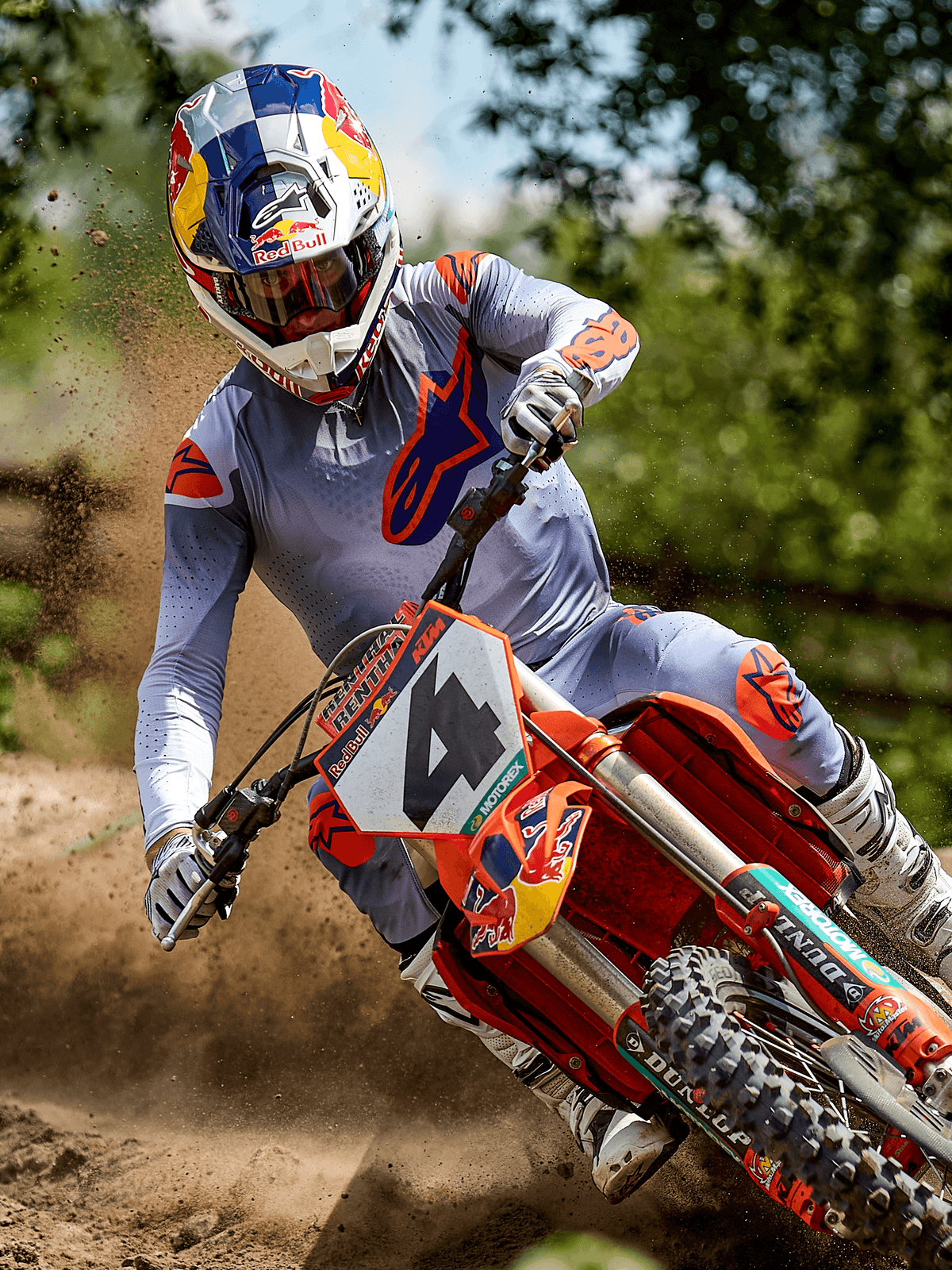 Motocross-Fahrer in Aktion mit dem Alpinestars 2025 Supertech Lipan Ausrüstungsset in Grau Orange Fluo, bestehend aus einem hellgrauen Trikot und einer Hose mit leuchtend orangefarbenen Astars-Logos, weißen Stiefeln und einem Red Bull-Helm auf einem orangefarbenen KTM-Motorrad auf einer Dirt-Strecke.