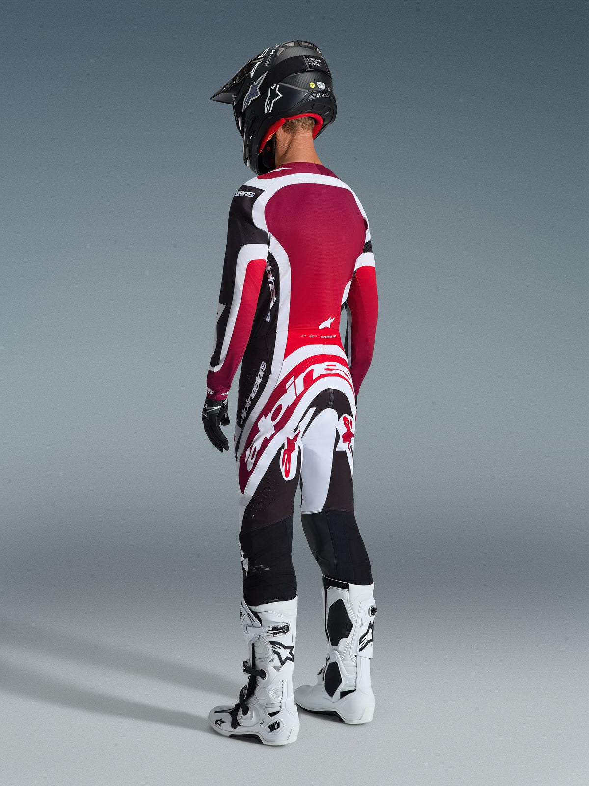 Motocrossrijder van achteren gezien die de 2026 Supertech AFD Vista Jersey draagt in zwart, rood en wit, bijpassende motorcrossbroek met opvallende witte Alpinestars-branding, zwarte helm en wit-zwarte laarzen, professionele off-road race-uitrusting
