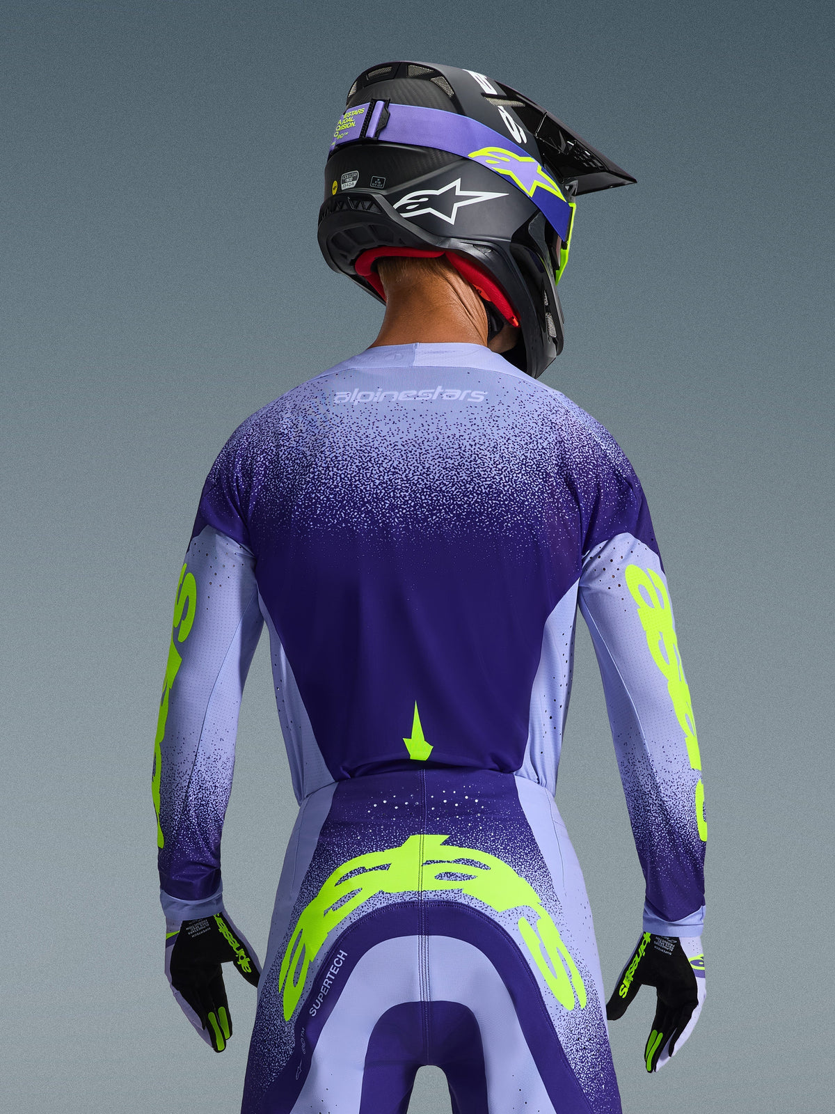 Alpinestars 2026 Supertech Scenz Jersey in Lila und Hellblau mit fluoreszierenden gelben Akzenten, Rückansicht am Fahrer mit passender Motocross-Hose, schwarzem Helm und Handschuhen, Hochleistungs-Sportausrüstung für den Offroad-Rennsport
