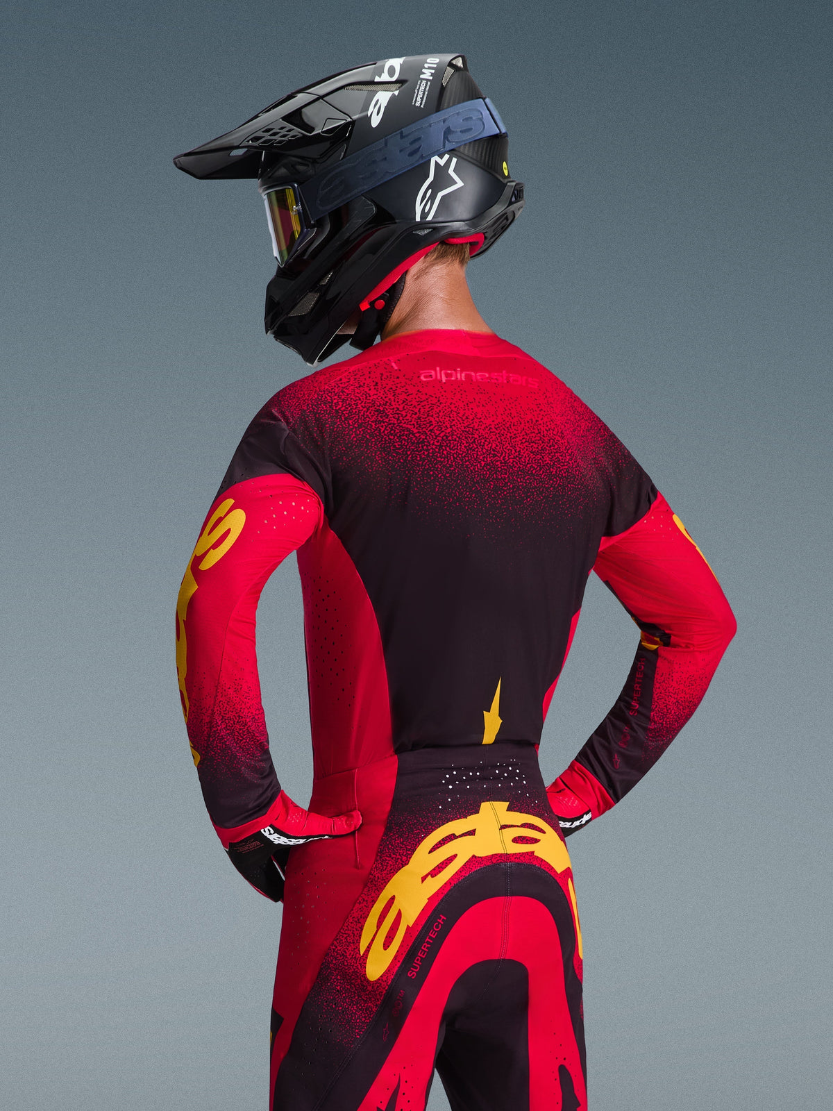 Maillot Alpinestars Supertech Scenz 2026, rouge, noir et jaune, vue arrière d'un pilote de motocross portant une tenue de course assortie comprenant un pantalon tout-terrain et un casque noir, présentant un design en dégradé rouge et noir avec un logo Alpinestars jaune vif