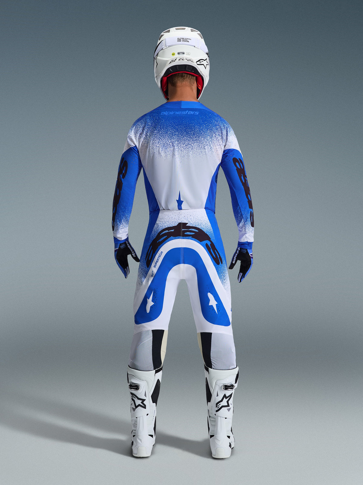 Pilote de motocross de dos portant l'ensemble Alpinestars 2026 Supertech Scenz en Ucla Blue White, maillot bleu et blanc avec logos noirs, pantalon assorti, casque blanc et bottes blanches, tenue de course de niveau professionnel pour le motocross tout-terrain