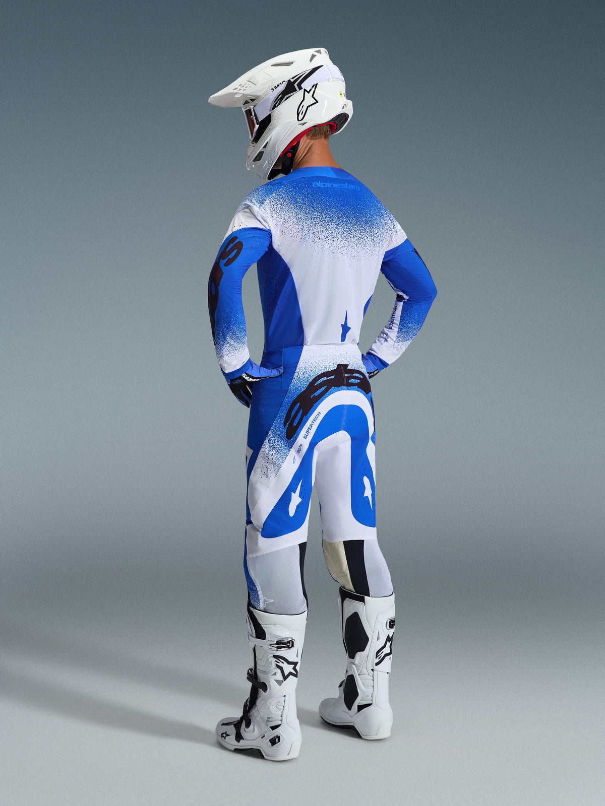Ensemble d'équipement Alpinestars Supertech Scenz 2026, vue arrière d'un pilote portant le maillot et le pantalon Ucla Blue White, casque de motocross blanc et bottes de course blanches, tenue de performance de course de niveau professionnel