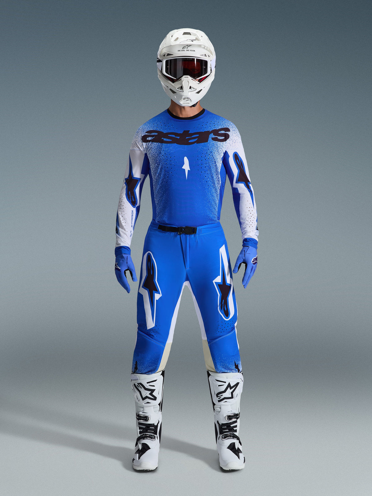 Pilote de motocross portant l'ensemble d'équipement Alpinestars Supertech Scenz 2026 en bleu Ucla et blanc, comprenant un maillot léger bleu et blanc avec logos noirs, un pantalon bleu assorti, un casque de course blanc et des bottes de motocross blanches, équipement de performance de niveau pro pour la course tout-terrain.