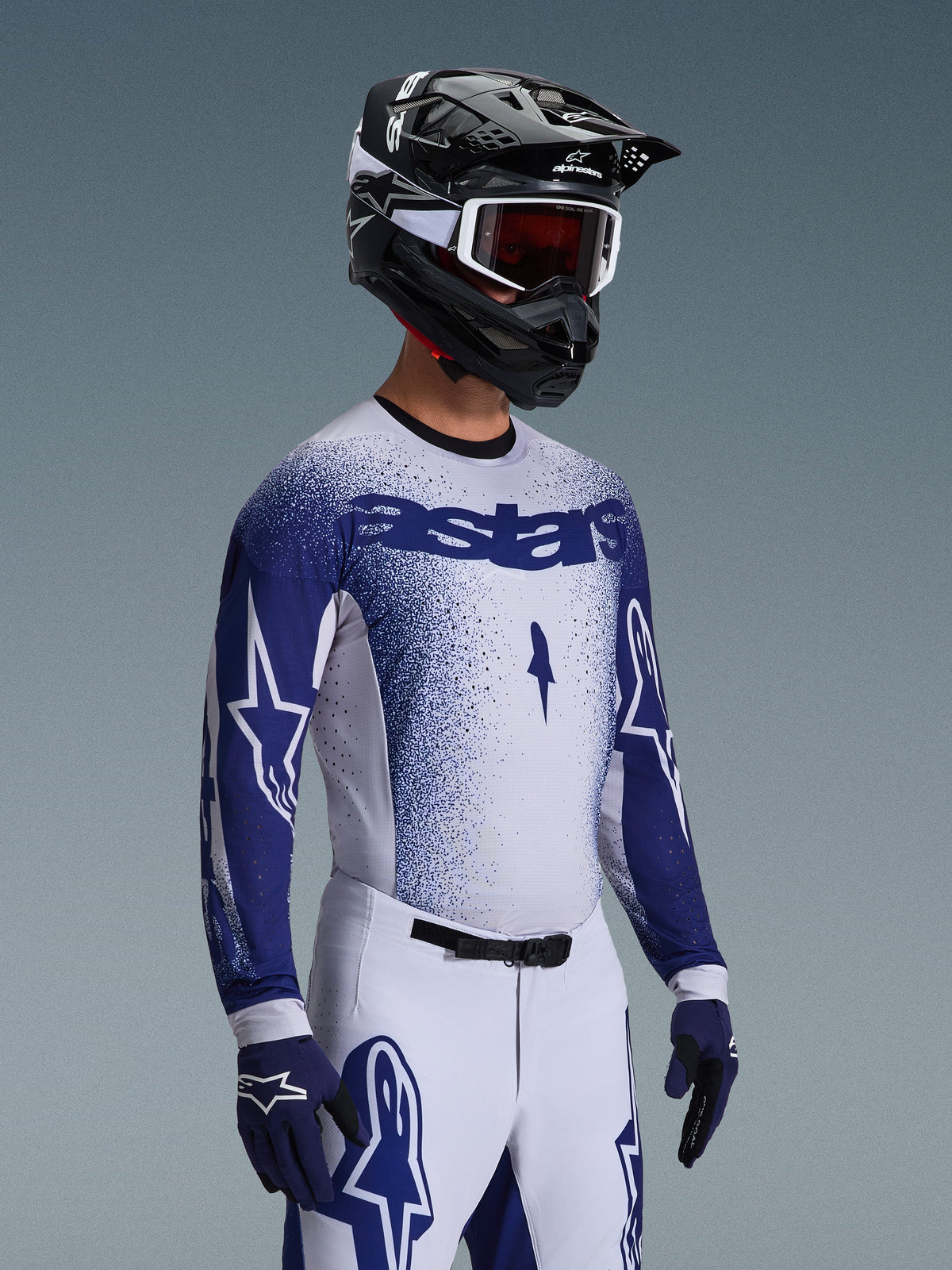 Pilote de motocross portant le Maillot Supertech Scenz 2026 d'Alpinestars en gris clair et bleu foncé, avec un pantalon de course blanc et bleu assorti, des gants bleus et un casque noir avec lunettes blanches. Le maillot présente un effet de dégradé bleu moucheté sur la poitrine et les manches, une coupe athlétique professionnelle et un logo 'astars' audacieux.
