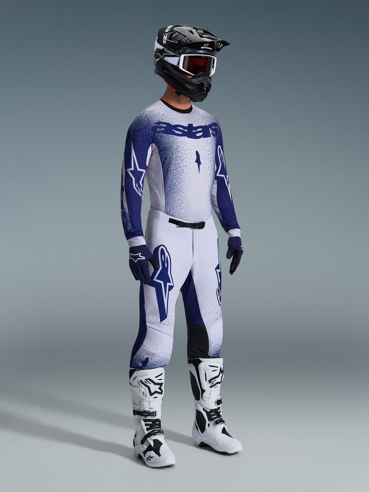 Pilote de motocross portant une tenue de course professionnelle complète, maillot Supertech Scenz 2026 en gris clair et bleu foncé, pantalon de motocross assorti avec graphismes d'étoiles, casque noir avec lunettes rouges, bottes de performance blanches et noires, gants bleus et blancs coordonnés pour la course tout-terrain