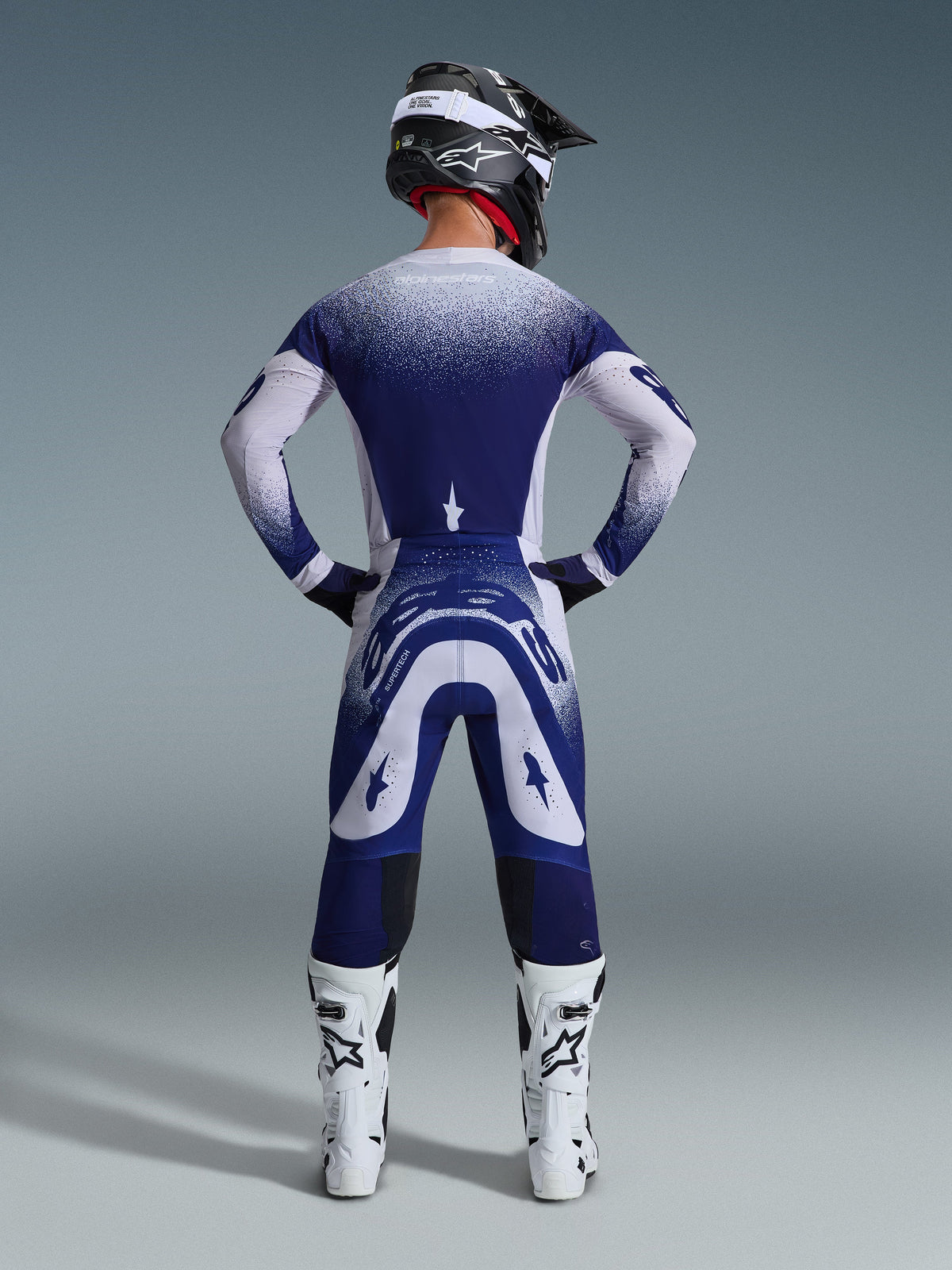 Pilote de motocross de dos, maillot Alpinestars Supertech Scenz 2026, gris clair et bleu foncé avec motif en dégradé pointillé, pantalon de motocross assorti, bottes de course blanches et casque noir, tenue complète de course tout-terrain