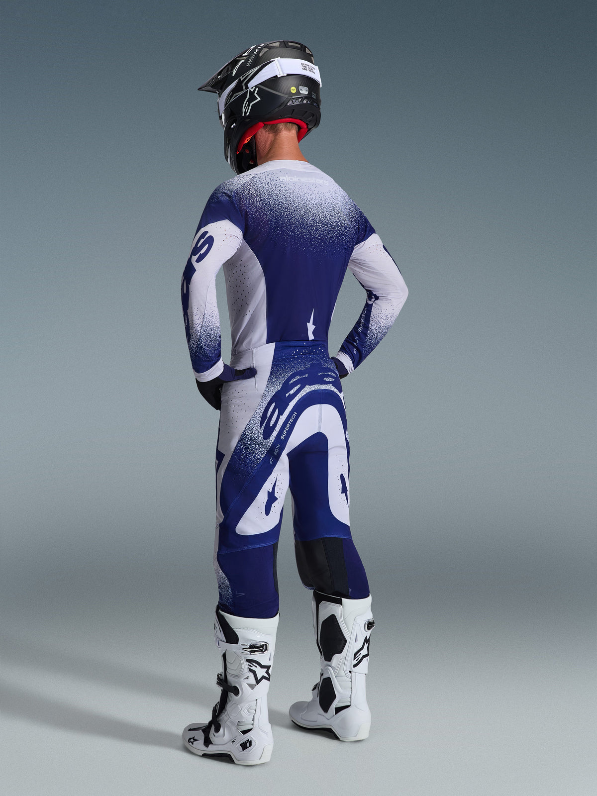 Vue arrière d'un pilote de motocross portant le maillot Alpinestars Supertech Scenz 2026 en gris clair et bleu foncé, avec un motif dégradé moucheté, coordonné à un pantalon assorti et des bottes de motocross blanches, équipement de course athlétique professionnel.