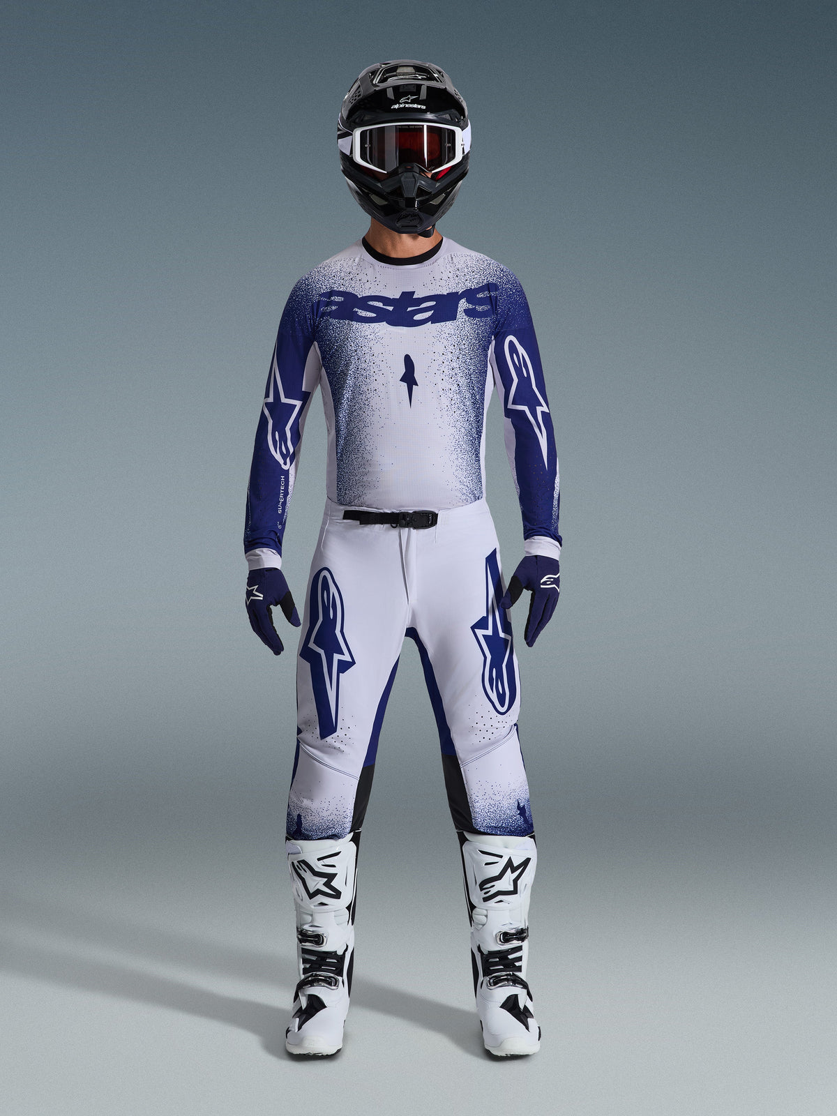 Pilote de motocross portant une tenue de course complète, Maillot Supertech Scenz 2026 en gris clair et bleu foncé, pantalon assorti gris clair avec logos bleus, casque noir et blanc avec lunettes rouges, bottes blanches et noires, gants bleu foncé, tenue coordonnée pour la course tout-terrain