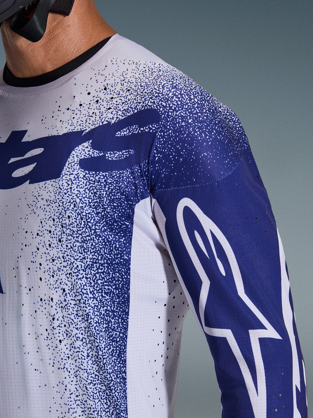 Gros plan sur le maillot Alpinestars Supertech Scenz 2026 en gris clair et bleu foncé, présentant des perforations au laser, un motif graphique moucheté et le logo Astars blanc sur la manche, coupe de performance motocross professionnelle