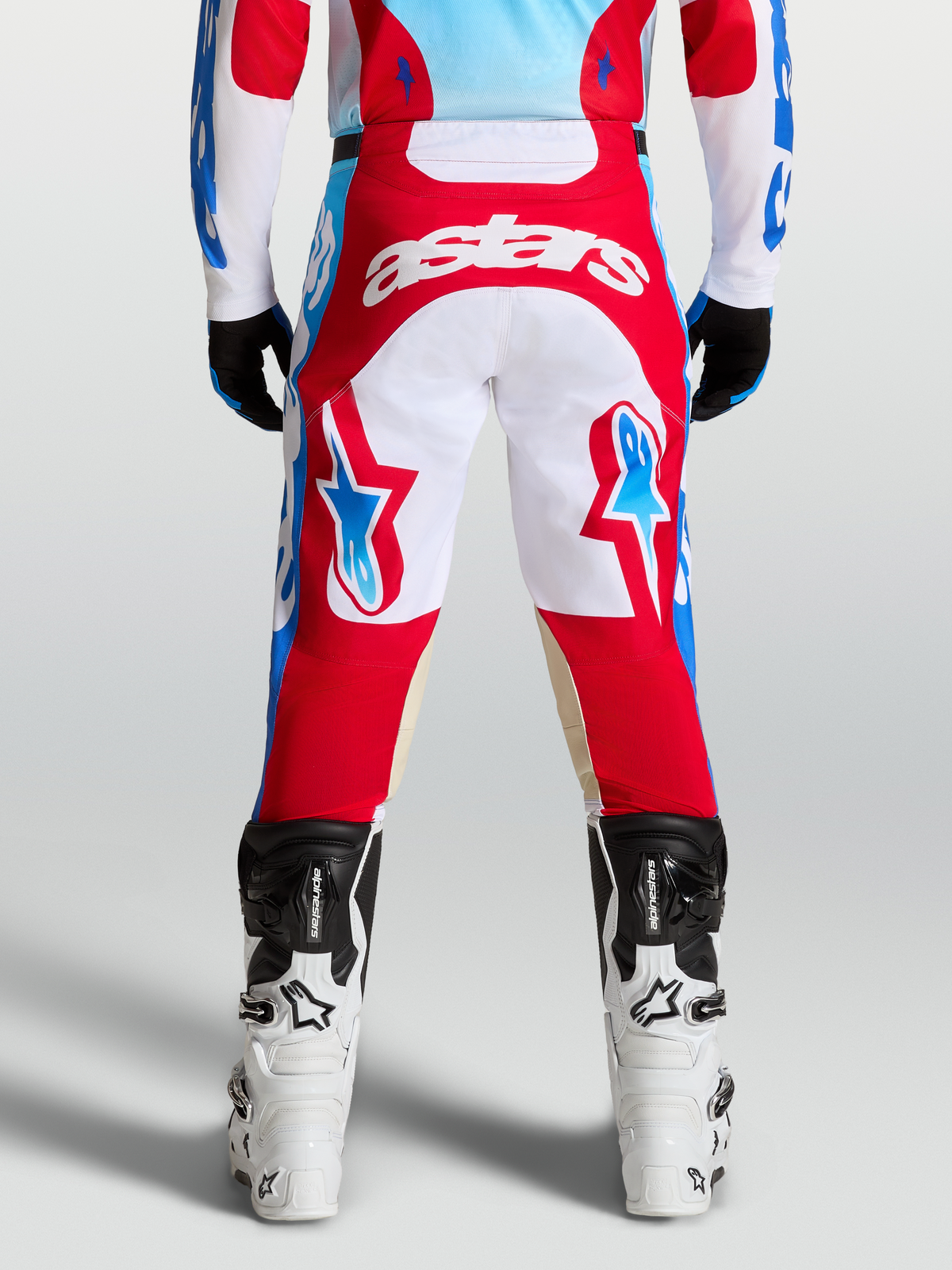 Alpinestars Pantalons Racer Hunt, pantalon de motocross tout-terrain pour homme, rouge et blanc avec accents bleus, vue de dos sur un pilote, construction ergonomique pré-courbée et tissu résistant