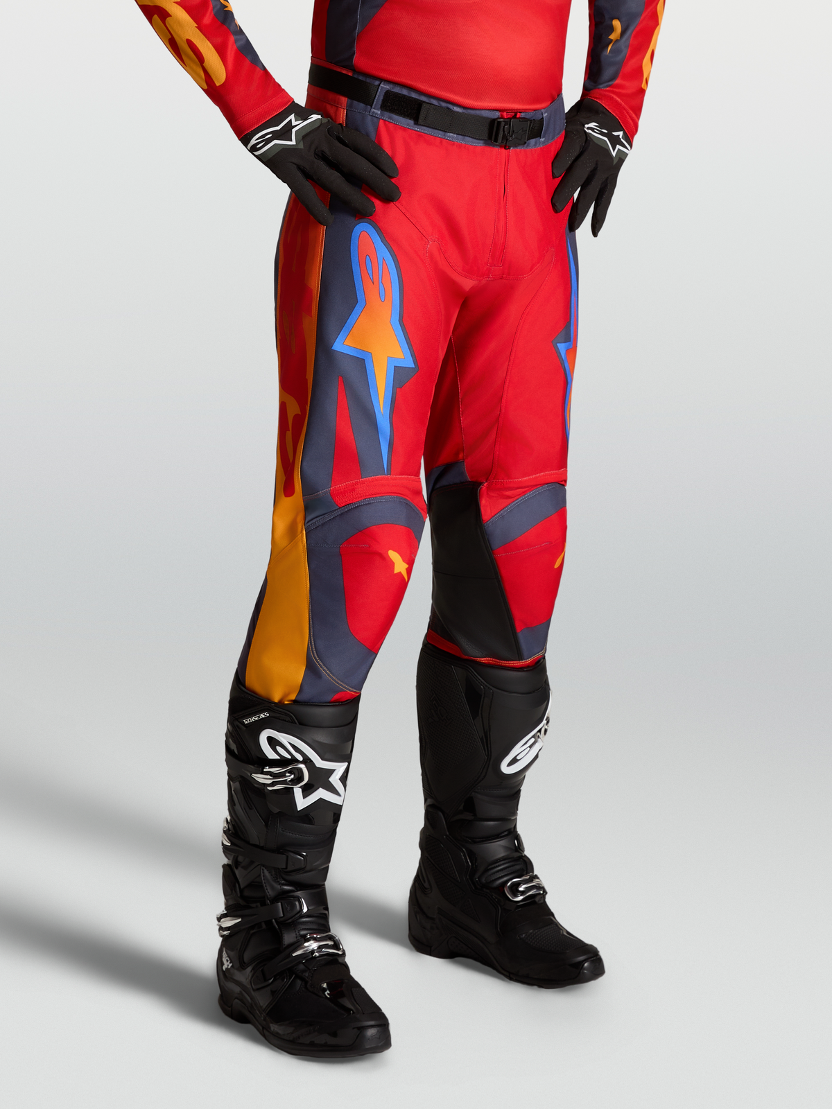 Alpinestars Pantalon Racer Hunt, pantalon de motocross pour homme rouge avec accents gris et corail, porté avec des bottes noires.