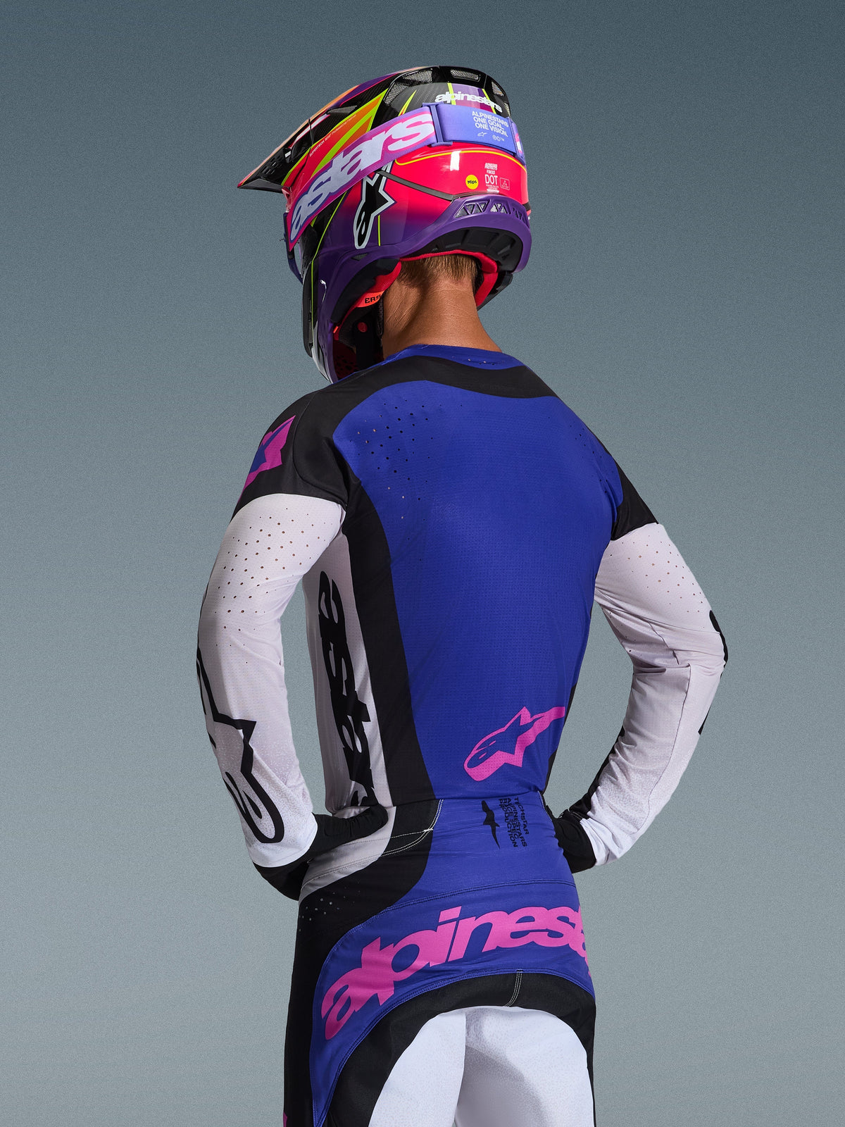 Motocross-Fahrer in der Rückansicht mit dem 2026 Techstar Knif Jersey, Schwarz, Hellgrau und Lila, mit weißen perforierten Ärmeln und pinken Alpinestars-Logos, kombiniert mit einem passenden lila-pinken Helm, professionelle Offroad-Rennausrüstung.