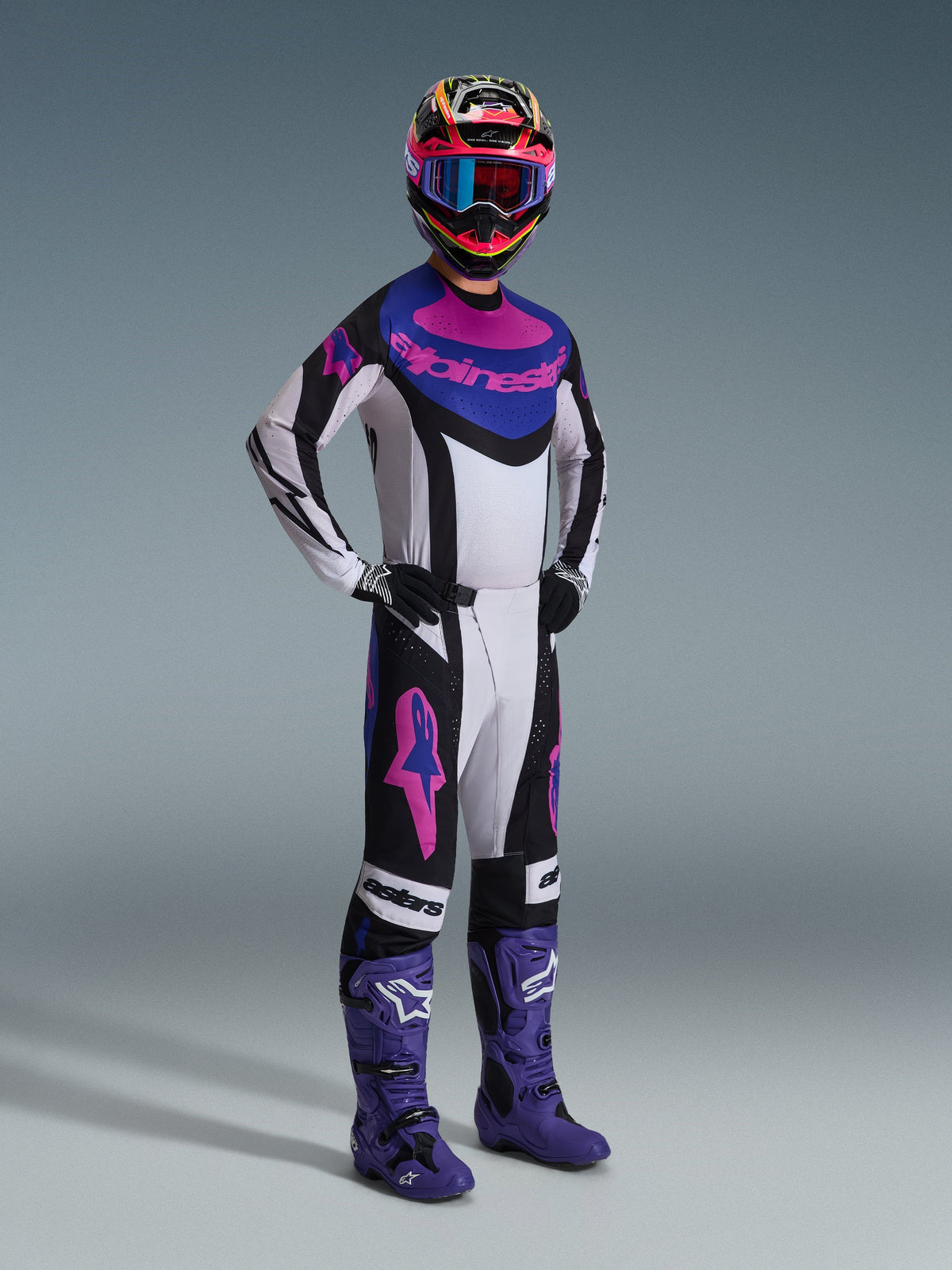 Motocross-Fahrer im Alpinestars 2026 Techstar Knif Jersey in Black Light Gray Purple, passende Hose mit großen pinkfarbenen Logos, lilafarbene Stiefel und ein mehrfarbiger Helm mit blauer Brille, komplette Rennausrüstung für Offroad-Performance