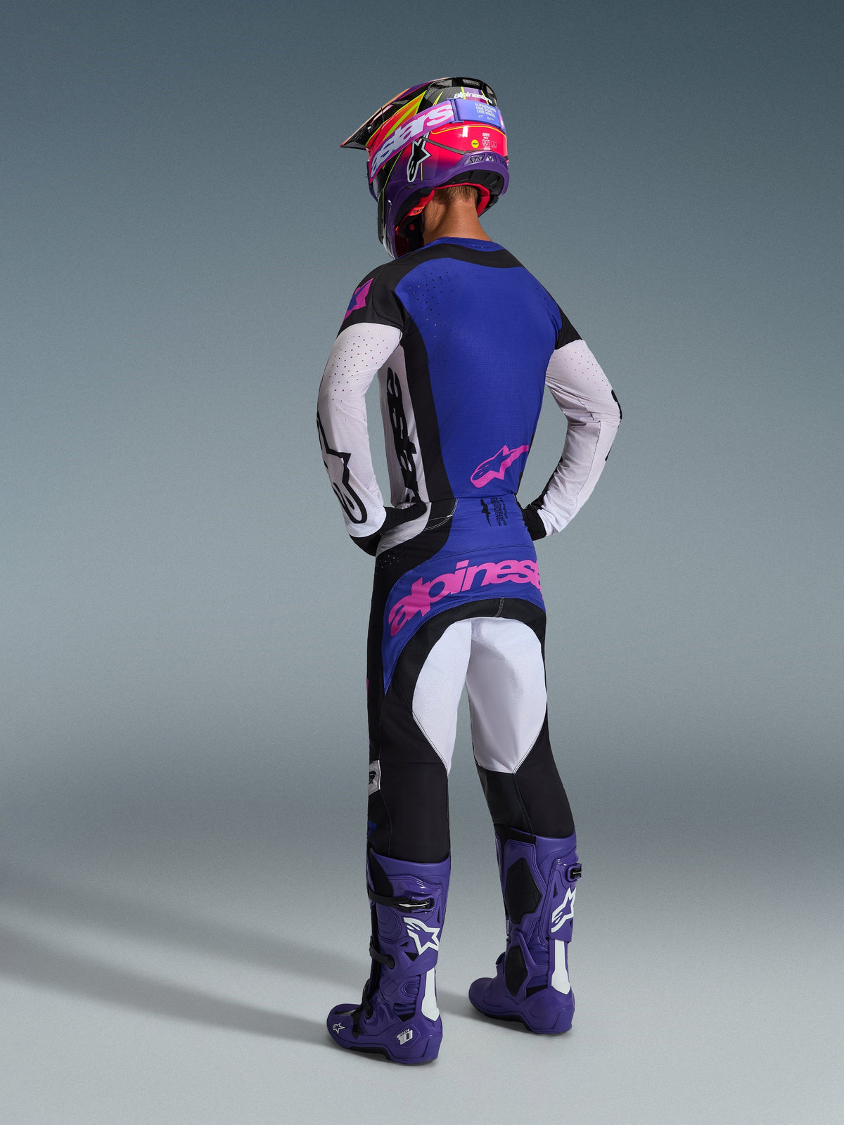 Motocross-Fahrer von hinten in kompletter Rennausrüstung, mit dem 2026 Techstar Knif Jersey mit blauem Rücken und weißen perforierten Ärmeln, passender schwarzer Hose mit pinkem Alpinestars-Branding, buntem Helm und lila Stiefeln, koordiniertes Outfit für Offroad-Wettbewerbe.
