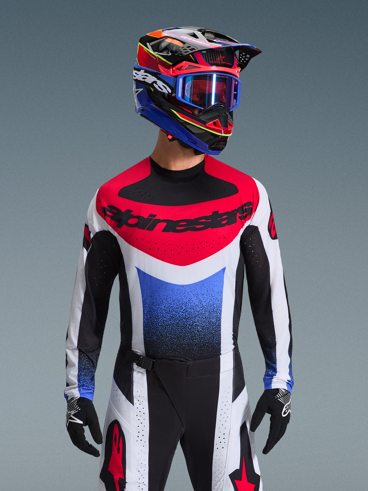 Ensemble Alpinestars 2026 Techstar Knif, pilote de motocross portant une tenue complète, maillot rouge, blanc et bleu avec logo noir imposant, pantalon noir et blanc assorti avec logo étoile rouge, casque multicolore avec lunettes bleues et gants noirs, tenue coordonnée pour la compétition tout-terrain