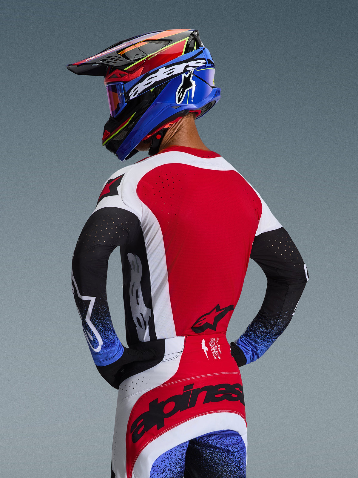 Pilote de motocross de dos portant la tenue Alpinestars Techstar Knif 2026, maillot rouge avec empiècements latéraux blancs et manches noires aux poignets mouchetés de bleu, pantalon rouge et blanc avec logo Alpinestars noir sur l'assise, casque bleu et rouge coordonné avec lunettes.