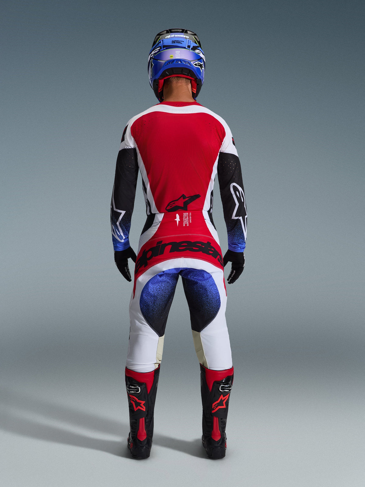 Pilote de motocross portant la tenue complète Alpinestars Techstar Knif 2026, vue de dos, comprenant un maillot rouge, blanc et noir avec des accents dégradés bleus sur les manches, un pantalon blanc et noir assorti avec un marquage rouge audacieux, un casque bleu et des bottes noires et rouges, tenue de course tout-terrain professionnelle.