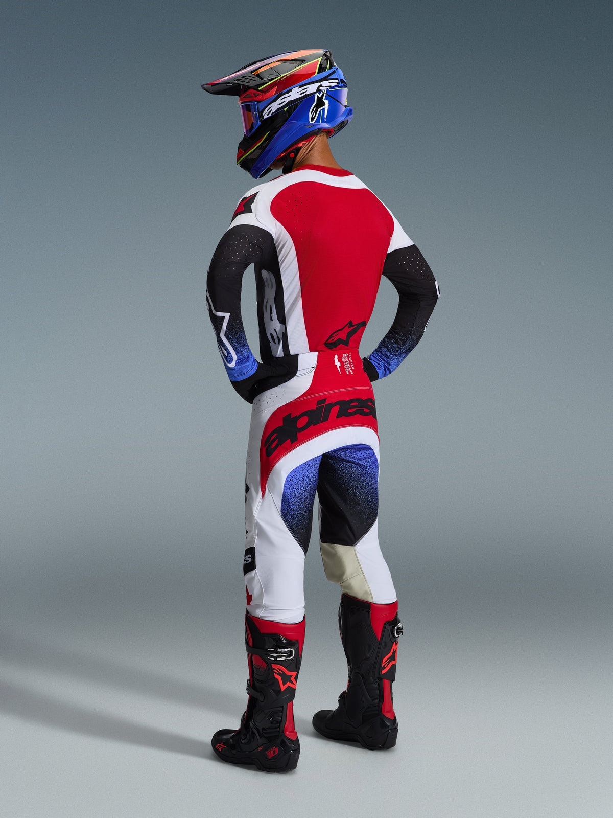 Pilote de motocross vu de dos portant la tenue complète Alpinestars Techstar Knif 2026, maillot noir, blanc et rouge avec manches à dégradé bleu et perforations laser, pantalon blanc et rouge assorti avec logo Alpinestars noir sur l'assise, casque bleu et noir avec lunettes rouges, bottes de motocross noires et rouges, équipement de course tout-terrain complet.